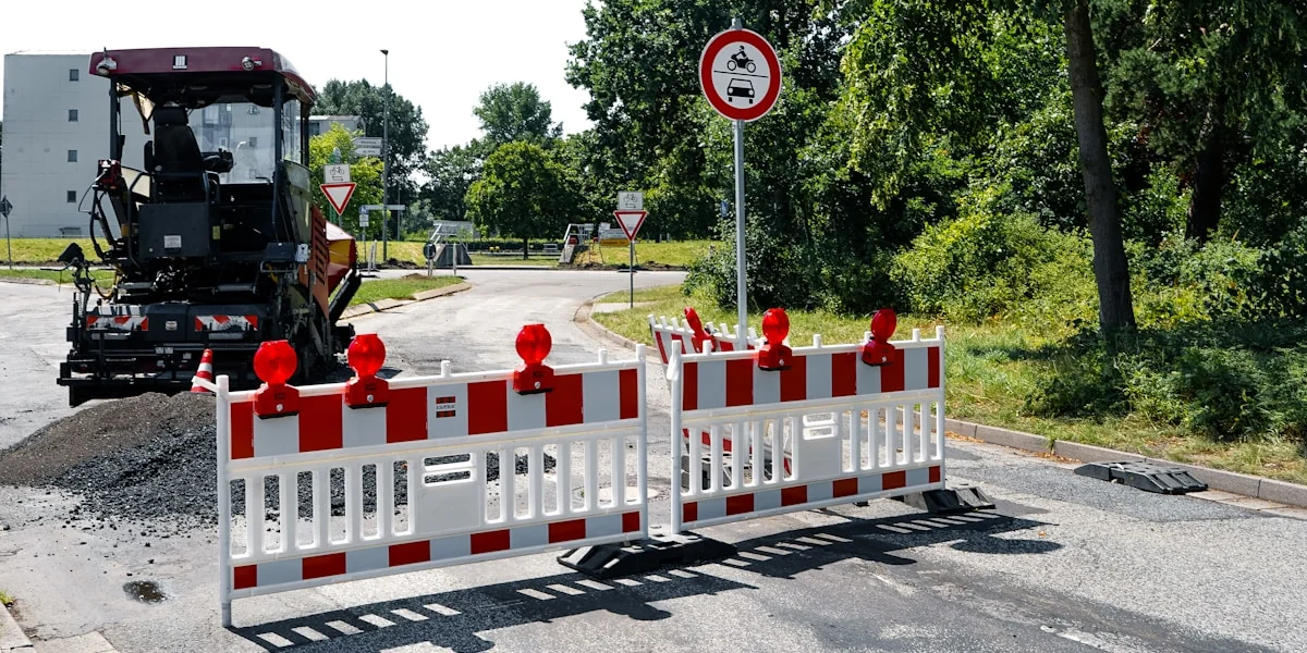 An excavator and road barriers block a construction site entrance.